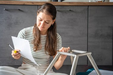 Sandalye monte ederken yerde oturmuş talimatları okuyan gülümseyen genç bir kadın. Dizinin devamı evde yapılan bağımsız çalışma neşe ve memnuniyeti gösteriyor. Yüksek kalite fotoğraf