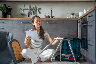 Sandalye monte ederken yerde oturmuş talimatları okuyan gülümseyen genç bir kadın. Dizinin devamı evde yapılan bağımsız çalışma neşe ve memnuniyeti gösteriyor. Yüksek kalite fotoğraf