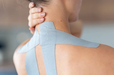 Kineziyoloji bandı boynuna ve omzuna uygulanan ve eliyle boynunu tutan genç bir kadının yakın görüntüsü. Modern rehabilitasyon yoluyla fizik tedavi ve kas iyileşmesi. 