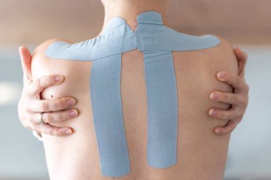 Sırtında ve omuzlarında kinezyoloji bandı olan genç bir kadın iki eliyle kendini kucaklıyor. Fizik terapi, vücut farkındalığı ve iyileşen dokunuşlarla duygusal benlik bakımı. Yüksek kalite fotoğraf