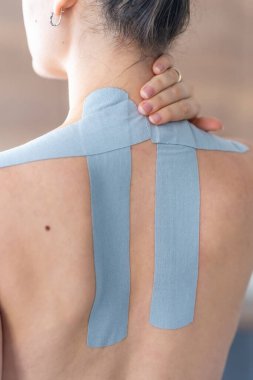 Kineziyoloji bandı boynuna ve omzuna uygulanan ve eliyle boynunu tutan genç bir kadının yakın görüntüsü. Modern rehabilitasyon yoluyla fizik tedavi ve kas iyileşmesi. 