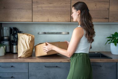Genç bir kadın mutfak tezgahında yemek pişirmek için önceden hazırlanmış malzemelerle dolu bir kutuyu açıyor. Yemek takımı teslimatı ve taze yemekle kolay ev yemeği kavramı.