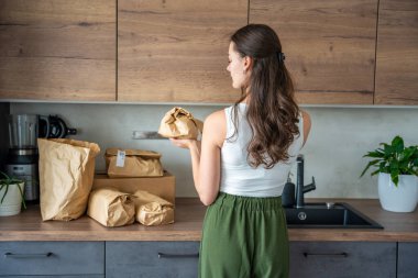 Modern mutfakta kahverengi bir kesekağıdının içinde yemek malzemeleri olan bir kadının arka görüntüsü. Kolay pişirme için önceden ölçülmüş market paketleriyle sürdürülebilir gıda teslimatı kavramı
