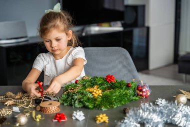Masada tek başına oturmuş Noel çelengi süsleyen küçük bir kız. Konforlu bir ev ortamında bağımsız yaratıcılık ve şenlikli bir ilişki anı