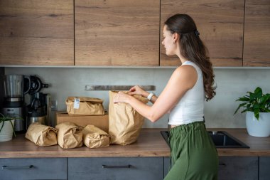 Modern bir mutfakta içinde yemek malzemeleri olan kağıt torbaları açan bir kadın. Ev yemeği için önceden ölçülmüş malzemelerle uygun market teslimatı kavramı