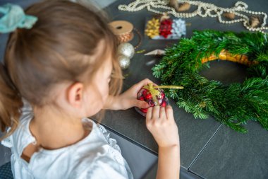 Küçük kız çelenk süslemeye başlamadan önce kutuları açıyor ve Noel süslerini hazırlıyor. Çocukların sezonluk geleneklere uyması için dikkatli bir tatil hazırlığı ve katılımı