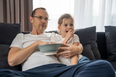Kanepede oturup televizyon izleyen ve patlamış mısır yiyen bir adam ve çocuk. Evde paylaşılan aile zamanı ve bağ..