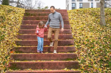 Düşen yapraklarla kaplı merdivenlerden inerken kızları tutan adam. Doğada ebeveyn-çocuk gezisi.