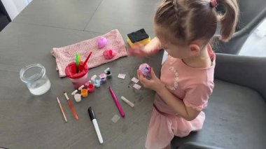 Küçük kız evde otururken Paskalya yumurtalarını renkli çıkartmalarla süslüyor. Yaratıcı Paskalya aktivitesi, basit yumurta süsleme tekniği ve kapalı alanda neşeli tatil hazırlığı.