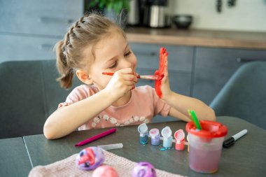 Paskalya yumurtalarını süslerken elini kırmızı boyayla boyayan küçük bir kız. Yaratıcı Paskalya aktivitesi, çocukluk merakı ve kapalı alanda eğlenceli tatil hazırlığı..