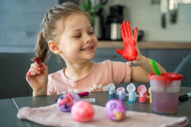 Paskalya yumurtalarını süslerken elini kırmızı boyayla boyayan küçük bir kız. Yaratıcı Paskalya aktivitesi, çocukluk merakı ve kapalı alanda eğlenceli tatil hazırlığı..