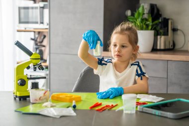 Mikroskop ve bilimsel aletlerle masada otururken cam kabın içinde büyüyen kristali inceleyen kız. Ev bilimi deneyi, merak ve kristal yetiştirme kiti yoluyla erken STEM eğitimi