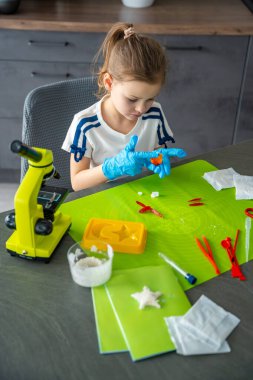Masada mikroskop ve laboratuvar aletleriyle kristal incelerken koruyucu eldiven giyen bir kız. Kristal büyüme kiti ile bilim deneyi ve erken kimya öğrenme