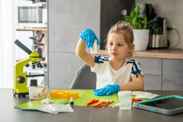 Mikroskop ve bilimsel aletlerle masada otururken cam kabın içinde büyüyen kristali inceleyen kız. Ev bilimi deneyi, merak ve kristal yetiştirme kiti yoluyla erken STEM eğitimi