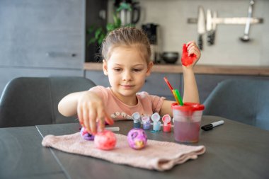 Paskalya yumurtalarını süslerken elini kırmızı boyayla boyayan küçük bir kız. Yaratıcı Paskalya aktivitesi, çocukluk merakı ve kapalı alanda eğlenceli tatil hazırlığı..