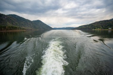 Teletskoye Gölü 'nden Altai Rusya' ya giden feribotun arkasında uyanma izleri var. Uzak kıyı şeridi ve dağları olan sakin bir su yüzeyi. Yüksek kalite fotoğraf