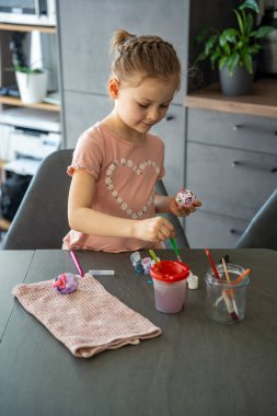 Renkli Paskalya yumurtasına ince fırçayla detaylar ekleyen odaklanmış bir çocuk. Sanatsal sabrı ve mevsimsel kutlamaları temsil eder. Mevsimsel yaratıcılık, odaklanma ve evde festival geleneği ruhu..