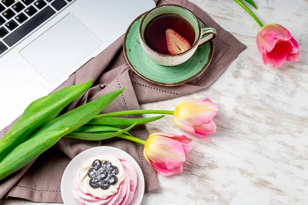 Egy csésze teát, tulipánt és egy tortát egy laptop mellett az asztalon. Kényelmes munka otthonról koncepció tavasszal — Stock Fotó