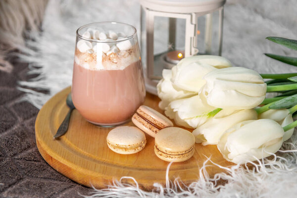 A glass of hot cocoa with marshmallows, macaroni cakes and white tulips on a tray in bed close up. Cozy spring home concept