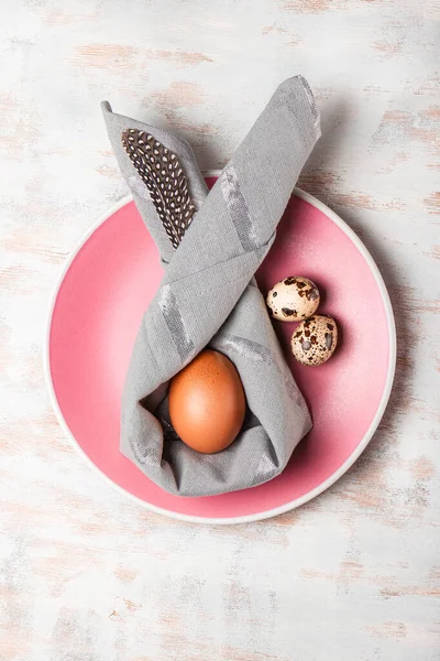  Peçeteden paskalya tavşanı ve pembe tabakta Paskalya yumurtası. Beyaz arka plan, üst manzara, dikey fotoğraf. Bayramlık Paskalya sofrası konsepti