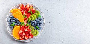 Çeşitli meyve ve böğürtlenlerden oluşan taze yaz salatasıyla internet afişi. Açık gri arka planda naneyle süslenmiş bir tabakta kivi, portakal, çilek ve yaban mersini, metin üst görünümü için boş alan