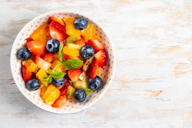Çeşitli meyve ve meyvelerden oluşan taze yaz salatası. Kivi, portakal, çilek ve böğürtlen, ahşap beyaz arka plan manzaralı nane ile süslenmiş bir tabakta.