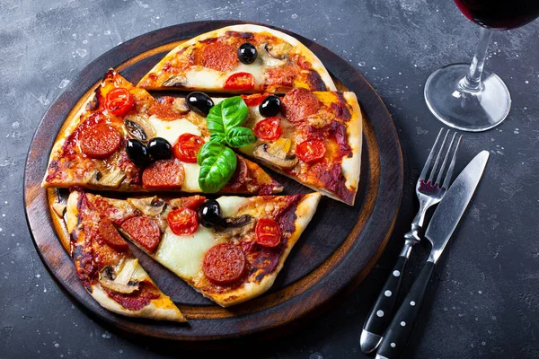 Pizza fatta in casa con mozzarella, salsicce al peperoncino, olive e basilico e un bicchiere di vino rosso in tavola — Foto Stock