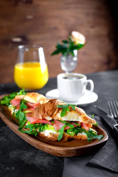 Croissanty s rukolou, avokádem a lososem, šálkem černé kávy a sklenicí pomerančové šťávy na stole, snídaňový koncept s měkkou zaostřenou svislou fotografií — Stock fotografie