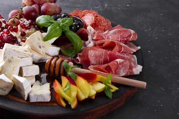  Şarap, jambon, pepperoni, peynir, üzüm, şeftali ve zeytin için ahşap bir tahtada aperatifler. Koyu gri arka planda abur cubur tahtası