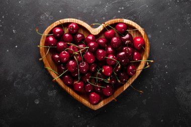 Koyu gri arka plan görünümlü, kalp şeklinde ahşap bir tabakta olgun kirazlar.