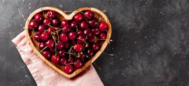 Koyu gri arka planda kalp şeklinde ahşap bir tabakta olgun kirazlar, metin için boş yeri olan web pankartı.