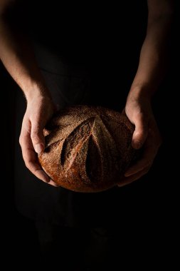 Fırıncının elindeki bir somun ekmeği siyah arka planda metin için boş alan, dikey karanlık ruh hali fotoğrafı. Yüksek kalite fotoğraf