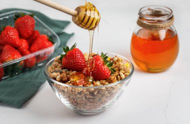 Beyaz arka planda izole edilmiş, cam bir tabakta çilek ve bal ile müsli. Bir bal çubuğu..