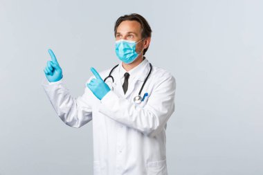 Covid-19, virüsü önleme, sağlık çalışanları ve aşı konsepti. Beyaz önlüklü orta yaşlı erkek doktor, tıbbi maske ve eldivenler sol üst köşeye bakıyor.