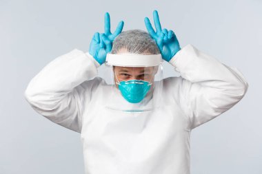 Covid-19, virüsü önleme, sağlık çalışanları ve aşı konsepti. Şahsi koruyucu ekipmanlarıyla mutlu iyimser doktor parmaklarını başının üstüne kaldırarak tavşan kulağı hareketi yapıyor.