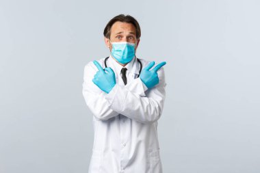 Covid-19, virüsü önleme, sağlık çalışanları ve aşı konsepti. Tıbbi maskeli, eldivenli, parmaklarını yana doğru gösteren kararsız ve kafası karışık bir doktor seçiminde yardıma ihtiyaç duyar.