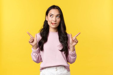 İnsan duyguları, yaşam tarzı ve moda kavramı. Rüya gibi stil sahibi Koreli kız, cazibe ve arzudan dudak ısırıyor, ilginç düşünceleri var, sol üst köşeye bakıyor, yan tarafları işaret ediyor, ürünü seçiyor.