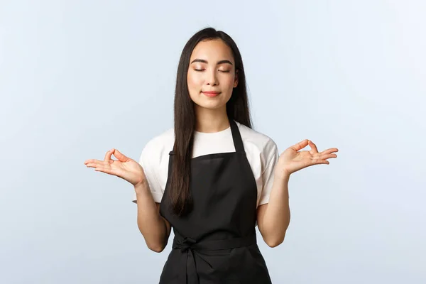 Coffee shop, small business and startup concept. Waitress being patient. Cute asian female barista in apron, close eyes and spread hands sideways mudra, zen gesture, meditating, do yoga at work
