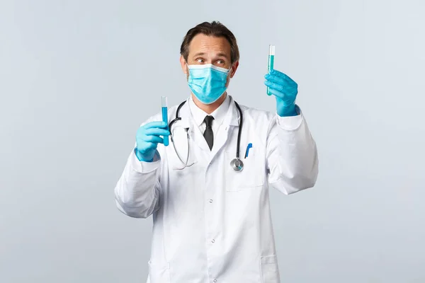 Covid-19: 바이러스 예방, 의료 종사자 및 예방 개념. 병원의 사려깊은 의사는 의료 마스크와 장갑을 착용하고 백신 샘플이 있는 두 개의 시험관을 바라본다 — 스톡 사진