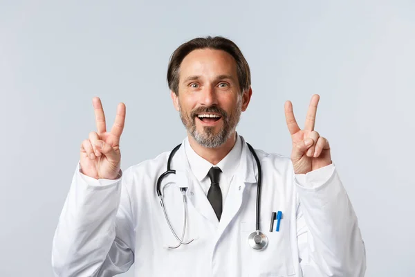 Covid-19, Prävention von Viren, Gesundheitspersonal und Impfkonzept. Aufgeregt lächelnde männliche Ärzte im weißen Mantel schauen begeistert, zeigen Frieden oder V-Zeichen mit erstaunter Miene — Stockfoto