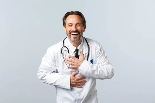 Covid-19, coronavirus outbreak, healthcare workers and pandemic concept. Carefree male doctor in white coat laughing out loud, sincere chuckle as having fun, hear good joke — Stock Photo, Image