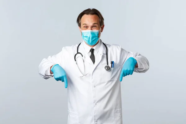 Covid-19: 바이러스 예방, 의료 종사자 및 예방 개념. 미디엄 마스크와 장갑을 낀 열정적 이고 잘생긴 남자 의사, 광고에 손가락을 아래로 지적 백인 배경 — 스톡 사진