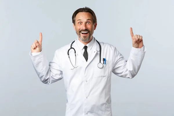 Covid-19, ξέσπασμα του ιού της στέψης, επαγγελματίες υγείας και πανδημία. Ενθουσιασμένος άντρας γιατρός με λευκό παλτό να δείχνει τα δάχτυλα ψηλά χαμογελώντας ενθουσιασμένος, λέγοντας υπέροχα νέα. — Φωτογραφία Αρχείου
