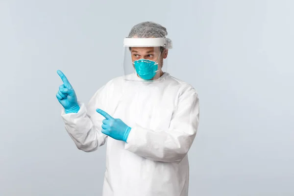 stock image Covid-19, preventing virus, healthcare workers and vaccination concept. Intrigued and surprised doctor in personal protective equipment raise eyebrows wondered, pointing fingers upper left corner