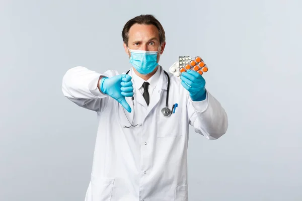 Covid-19, prévention du virus, personnel de santé et concept de vaccination. Médecin sérieux mécontent dans le masque médical et les gants, montrer pouce vers le bas et les médicaments, pilules de mauvaise qualité, mauvaise prescription — Photo