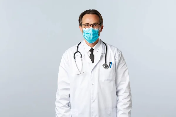 Covid-19, preventing virus, healthcare workers and vaccination concept. Serious confident male doctor in white coat and medical mask, wearing glasses, ready treat patient with influenza — Stock Photo, Image