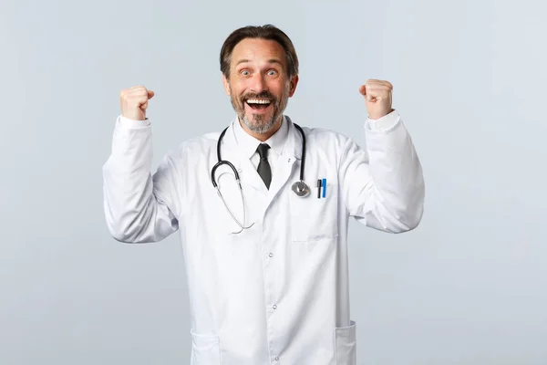 Covid-19, ξέσπασμα του ιού της στέψης, επαγγελματίες υγείας και πανδημία. Ανακουφισμένος ευτυχισμένος άνδρας γιατρός λαμβάνει καλά νέα, σηκώνοντας τα χέρια ψηλά στη γιορτή και χαμογελώντας ικανοποιημένος, θριαμβεύοντας — Φωτογραφία Αρχείου