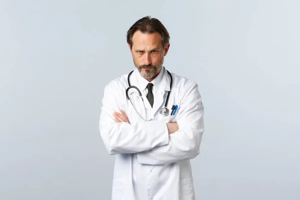 Covid-19, coronavirus outbreak, 의료 종사자와 전염병 컨셉트. 심각하게 실망 한 흰 코트를 입은 남자 의사, 양팔 가슴을 교차하는 모습 그리고 이마의 판 단 아래서 바라봄 — 스톡 사진