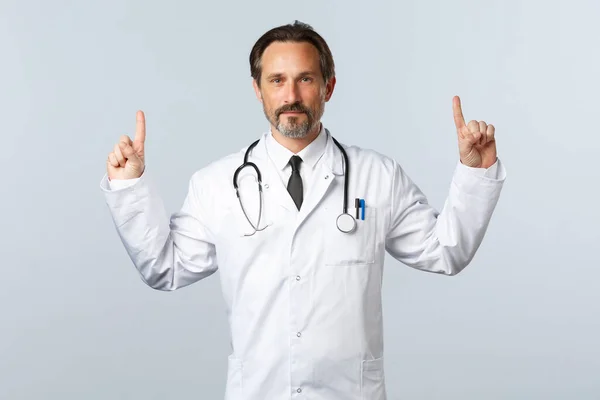 Covid-19, utbrott av coronavirus, vårdpersonal och pandemikoncept. Självsäker vuxen läkare i vit rock, stetoskop, pekar finger upp och ler, ordinera piller, reklam produkt — Stockfoto