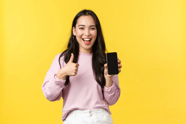 People emotions, lifestyle leisure and beauty concept. Smiling asian woman 20s, showing smartphone display, recommend application or mobile game, showing thumbs-up in approval, yellow background — Stock Photo, Image